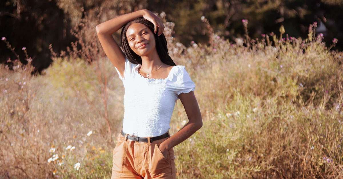 Leah Thomas stands in a field of tall grass and wild flowers, right arm resting easily overhead and left hand tucked into her pocket. The photo is warm, with golden-hour colors and a soft, bokeh heavy background.