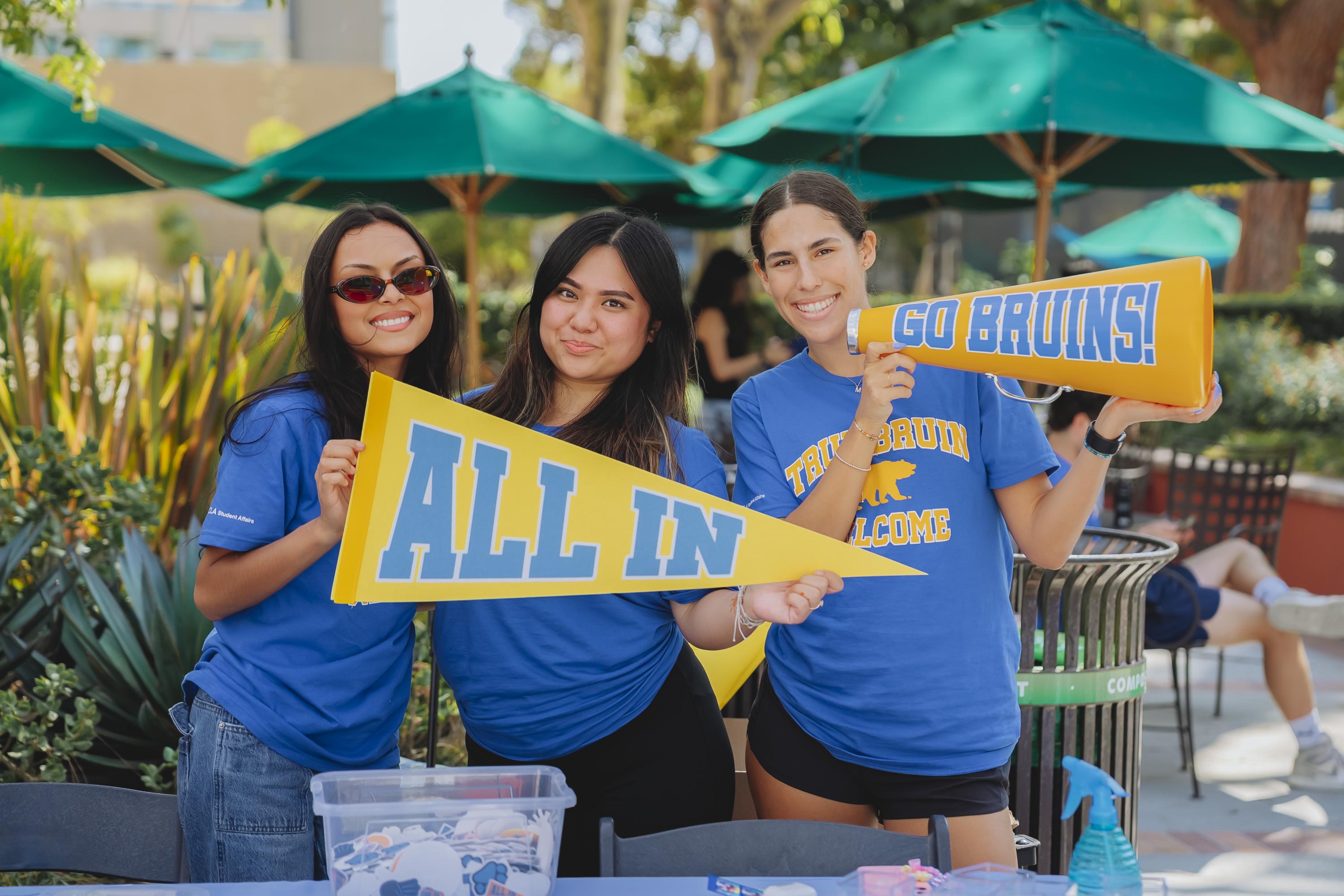 Image of three students smiling holding a "all in" pennant and a "go bruins" megaphone